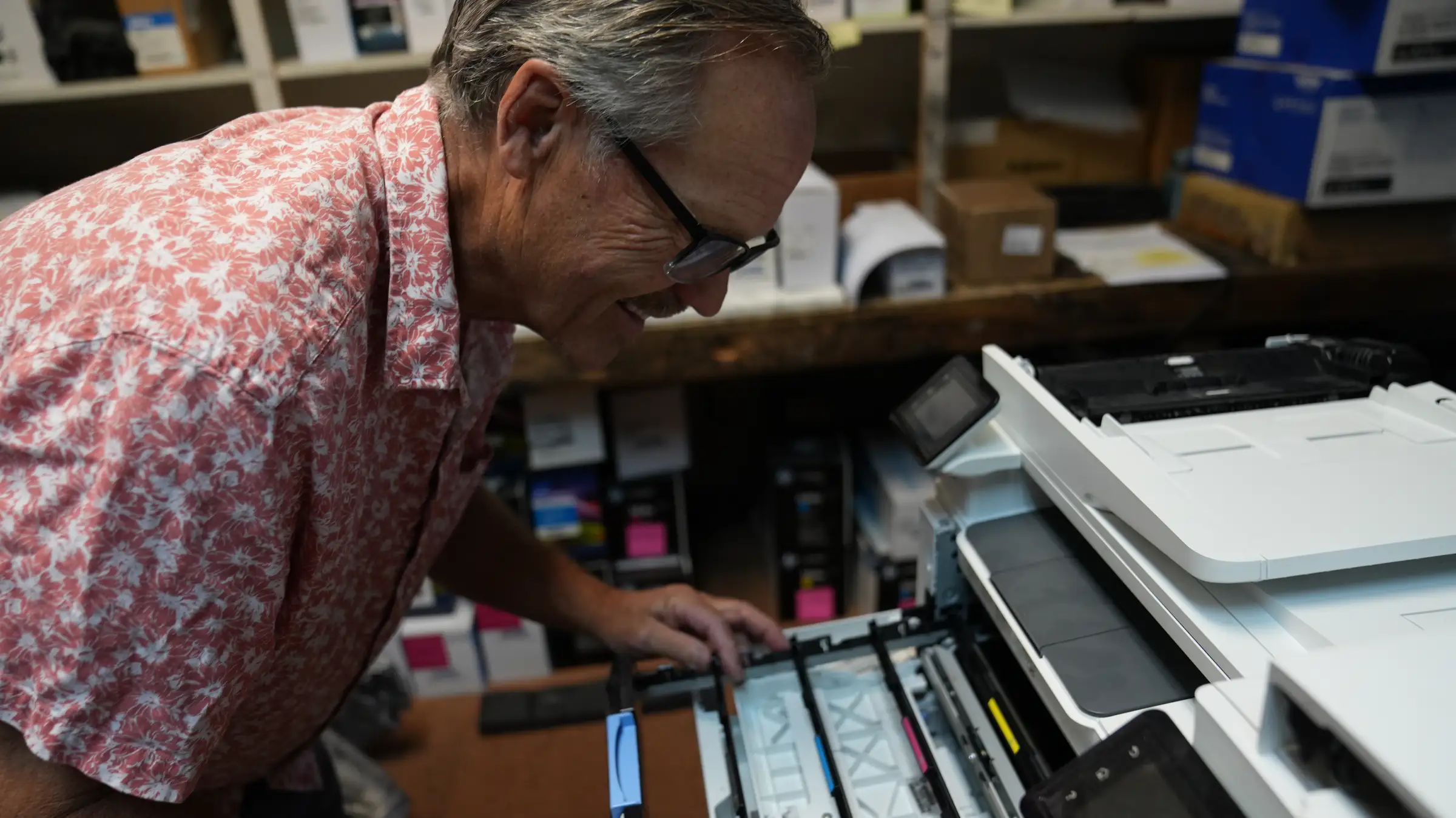 George Simmons, founder of GMS Lazerworks, smiling as he works inside an open color laser printer at the Novato shop